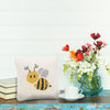 Bee knitted gift pillow with a neutral background and an adorable honeybee design on its front on a table next to flowers, books, and a coffee cup