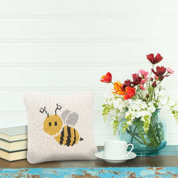 Bee knitted gift pillow with a neutral background and an adorable honeybee design on its front on a table next to flowers, books, and a coffee cup