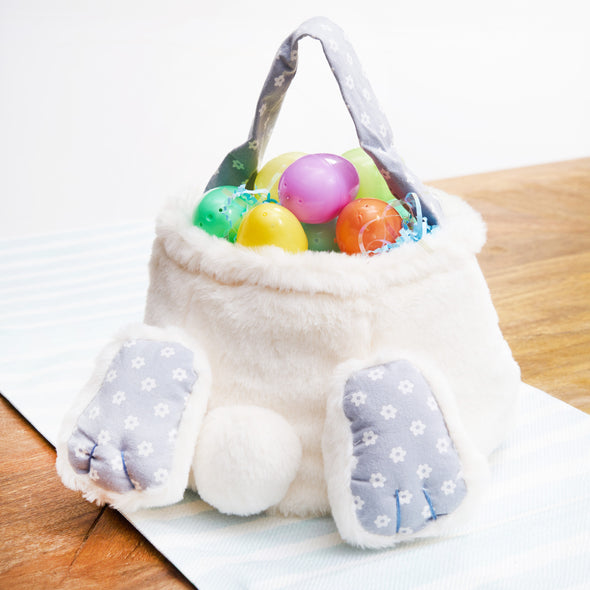 cotton tail bunny basket with two paws and a fluffy bunny tail. the handle and paws are light blue and have delicate white flowers. the basket is filled with easter eggs on a table
