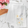 flour sack towel with three classic daisy flowers and two dragonflies swarming the flowers on a marble counter