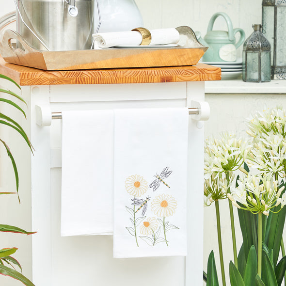 flour sack towel with three classic daisy flowers and two dragonflies swarming the flowers hanging from a counter