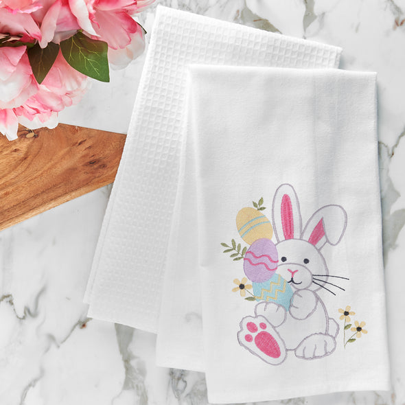 flour sack towel with an adorable bunny holding decorated easter eggs and surrounded by delicate flowers on a marble counter