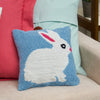 The Easter Bunny hooked pillow features a white rabbit against a blue background with a pom-pom tail on a couch with a pink and light blue pillow