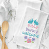 flour sack towel with the phrase every bunny welcome in bright spring colors. two bunnies sit on top of the phrase while a group of flowers are below. this towel is on a marble counter