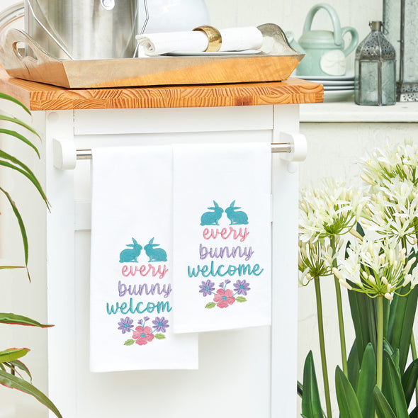 flour sack towel with the phrase every bunny welcome in bright spring colors. two bunnies sit on top of the phrase while a group of flowers are below. two of these towels hang from a counter