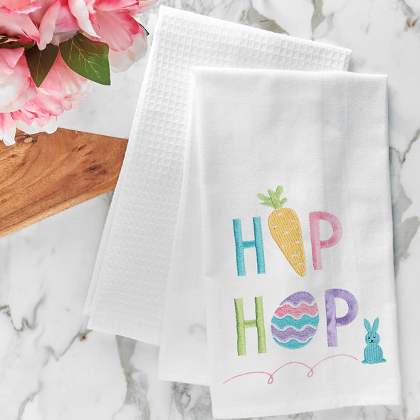 a flour sack towel with the words hip hop in pastel Easter hues. The I in Hip is replaced by a carrot and the O in Hop is replaced by an Easter egg. A bouncing bunny sits next to this phrase. this towel is on a marble countertop