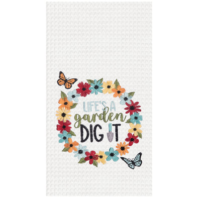 embroidered waffle weave towel with a wreath of colorful flowers surrounding the phrase Life's a Garden Dig It with a shovel creating the I. Butterflies flutter on the outside of the floral wreath.
