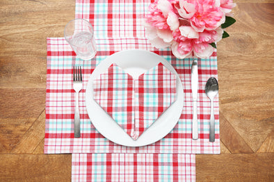 Red and light blue plaid napkin in a heart shape on a white place on a placemat and runner with the same pattern