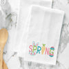 embroidered waffle weave towel with the word spring in pastel bright colors. bunny ears and paws stick out between the P, an open easter egg surrounds the G, and the I is a carrot. the towel is on a marble counter