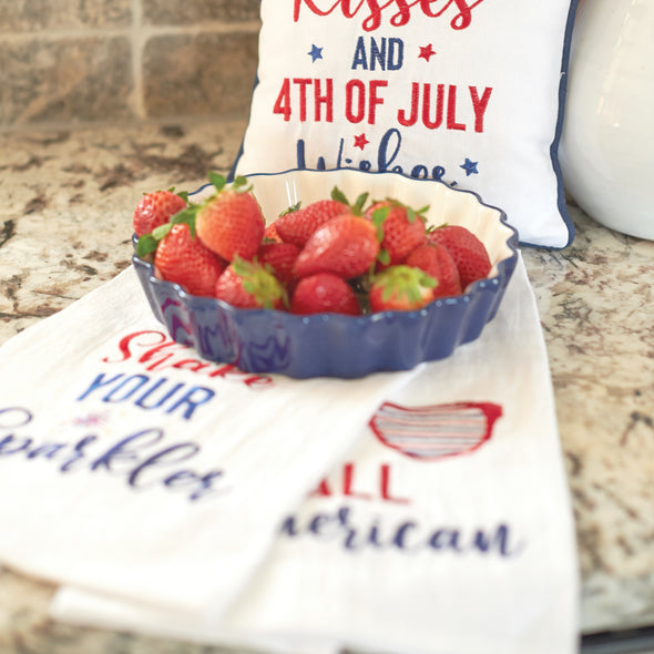 flour sack towel with the words all American paired with sunglasses that feature red white and blue stars and stripes paired with additional patriotic decor and a dish of strawberries