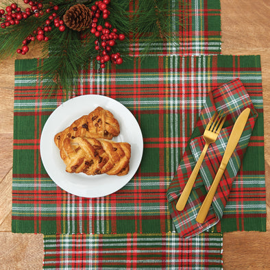 woven table linens with a forest green background and red, white, and yellow accents on a wood table surrounded by holly berries, pinecones, and holiday decor