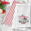 a waffle weave towel with a log cabin surrounded by the words Christmas in the woods in different scripts and decorated with holly berries and flowers on a marble countertop