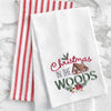 a waffle weave towel with a log cabin surrounded by the words Christmas in the woods in different scripts and decorated with holly berries and flowers on a marble countertop