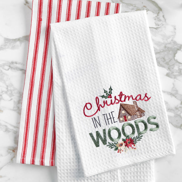 a waffle weave towel with a log cabin surrounded by the words Christmas in the woods in different scripts and decorated with holly berries and flowers on a marble countertop