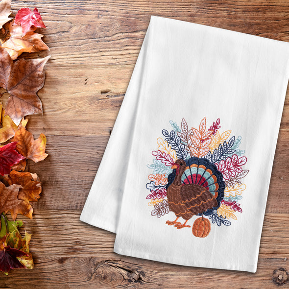 flour sack towel with detailed feathers spanning across the back of a turkey in a variety of fall colors standing next to a pumpkin on a wood table next to fall foliage