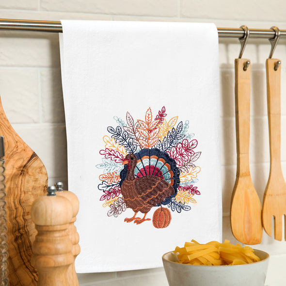 flour sack towel with detailed feathers spanning across the back of a turkey in a variety of fall colors standing next to a pumpkin hanging on a towel bar in the kitchen