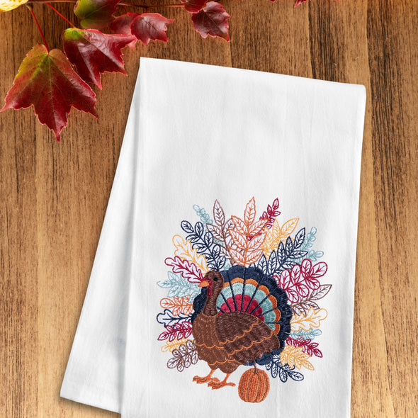 flour sack towel with detailed feathers spanning across the back of a turkey in a variety of fall colors standing next to a pumpkin on a wood table next to fall foliage