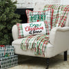 tufted and embroidered design with the phrase cozy christmas at home in a cursive font on a white background. the edges have a red green and white plaid pattern on a chair with a matching pillow and throw next to a Christmas tree and presents