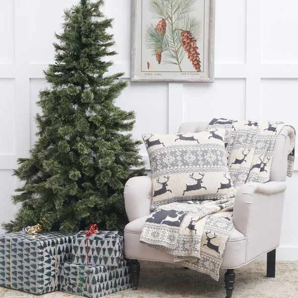 knitted pillow with a natural background and rows of leaping deer and snowflake patterns in light grey with a matching throw on a chair next to a Christmas tree and presents