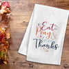 flour sack towel with the phrase Eat Pray & Give Thanks in inviting Autumn colors and a delicate script highlighted by flourishes on a wood table next to fall foliage