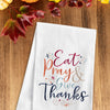 flour sack towel with the phrase Eat Pray & Give Thanks in inviting Autumn colors and a delicate script highlighted by flourishes on a wood table next to fall foliage