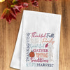 flour sack towel with harvest related words in different font styles arranged in different directions. decorative elements like pumpkins and foliage are added in the mix on a wood table with fall foliage