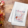 flour sack towel with harvest related words in different font styles arranged in different directions. decorative elements like pumpkins and foliage are added in the mix on a wood table with fall foliage