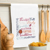 flour sack towel with harvest related words in different font styles arranged in different directions. decorative elements like pumpkins and foliage are added in the mix hanging from a kitchen towel bar