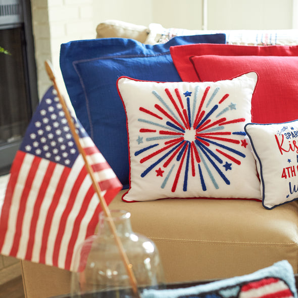 a tufted pillow featuring a bursting firework pattern of blue and red stripes paired with delicate stars. the pillow is trimmed in red and complemented by additional patriotic decor including pillows and a flag.