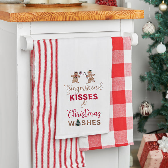 embroidered flour sack towel with the phrase gingerbread kisses and Christmas wishes below a gingerbread man and gingerbread woman hanging off a kitchen countertop