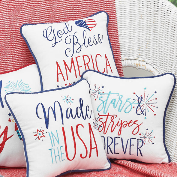 an embroidered pillow with the words God bless America in various font styles and underscored with tiny red and blue stars. a heart resembling the stars and stripes of the American flag complements the design. paired with additional patriotic gift pillows