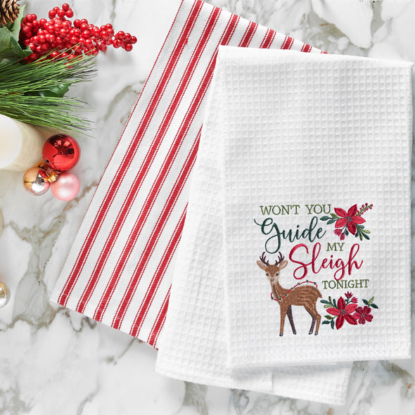 waffle weave towel with the phrase won't you guide my sleigh tonight in different font styles nestled between an adorable reindeer and red flower arrangements on a marble countertop