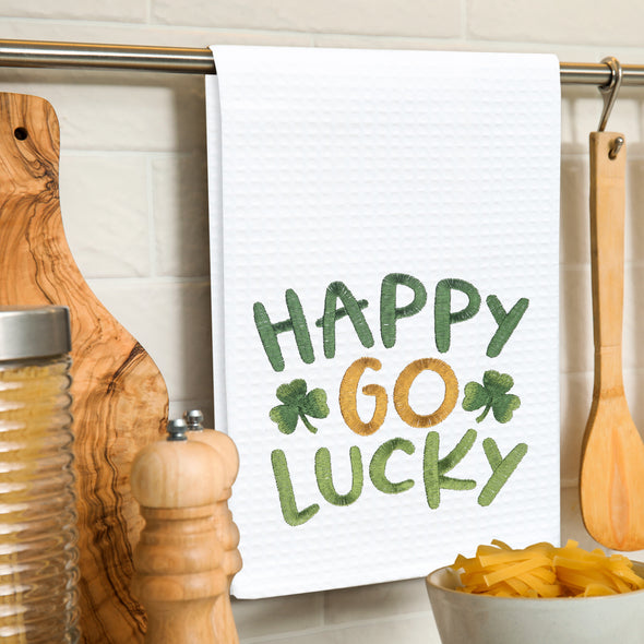 embroidered waffle weave towel with the words Happy Go Lucky surrounded by two clovers hanging on a kitchen towel rack