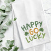 embroidered waffle weave towel with the words Happy Go Lucky surrounded by two clovers on a marble counter next to foliage