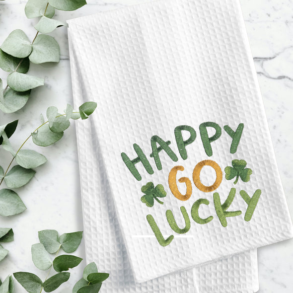 embroidered waffle weave towel with the words Happy Go Lucky surrounded by two clovers on a marble counter next to foliage