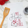 embroidered flour sack towel with the phrase my heart is full next to a letter bursting with pink hearts on a marble kitchen counter