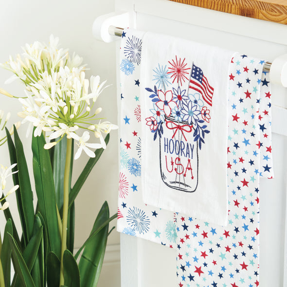 flour sack towel with a mason jar filled with flowers sparklers and an American flag with the words hooray USA hanging with other patriotic towels next to flowers