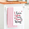 flour sack kitchen towel with an embroidered phrase of I love you more than wine surrounded by little hearts hanging from a kitchen counter