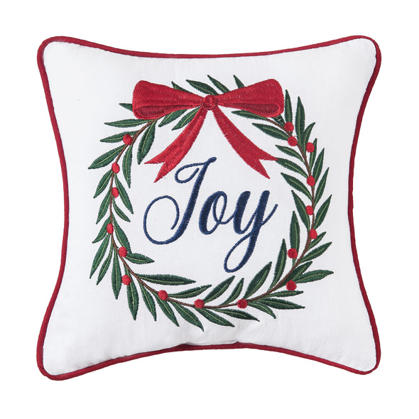 embroidered pillow with a white background trimmed in red. An abstract wreath sits in the center and topped with a red bow with the word Joy in the center.