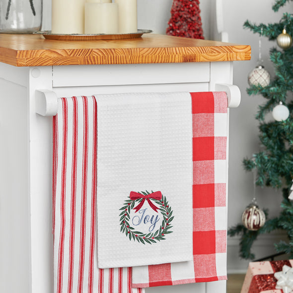 waffle weave towel with a wreath topped with a red bow and the word joy in the center hanging from a kitchen countertop