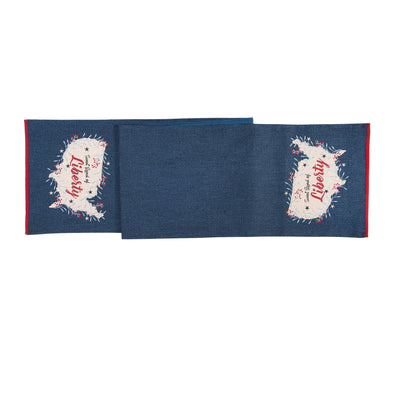 an applique and embroidered table runner with a denim blue background with each end featuring the shape of America embroidered on each end and the words Sweet Land of Liberty inside. The shape of America is outlined in a botanical and berry border.