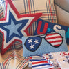 a hooked pillow featuring a light blue background and a pair of sunglasses with one side featuring blue with white stars and the other with red and white stripes to resemble the American flag surrounded by additional patriotic pillows and decor