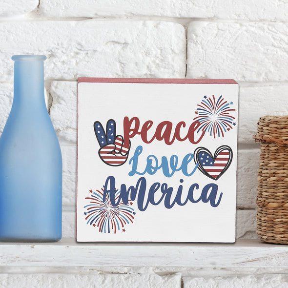 shelf block with the words peace love america surrounded by patriotic motifs on a shelf next to a blue bottle and against a brick wall