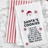 embroidered flour sack towel with instructions to make Santa's cookies including ingredients like love, joy, and sugar below a picture of Santa's face and surrounded by red snowflakes on a marble countertop