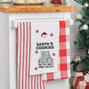 embroidered flour sack towel with instructions to make Santa's cookies including ingredients like love, joy, and sugar below a picture of Santa's face and surrounded by red snowflakes hanging from a kitchen countertop