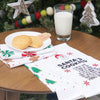 embroidered flour sack towel with instructions to make Santa's cookies including ingredients like love, joy, and sugar below a picture of Santa's face and surrounded by red snowflakes hanging from a kitchen countertop on a wood table in front of a decorated Christmas tree