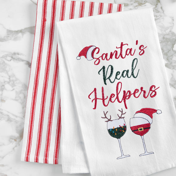 embroidered flour sack towel with the phrase Santa's real helpers above two glasses of holiday decorated wine on a marble countertop