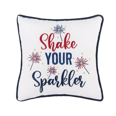 a white embroidered pillow with flourishing text that reads shake your sparkler surrounded by bursts of bright sparklers and tiny yellow stars