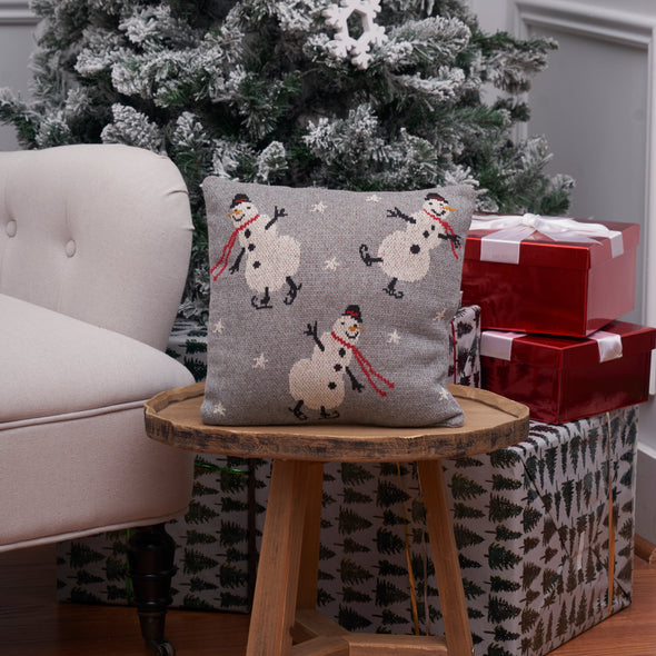 grey knitted pillow with snowmen in ice skates and red scarfs on a wood table next to a Christmas tree and presents