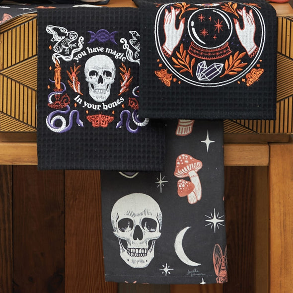 black kitchen towels with mystical motifs including skulls, potion bottles, snakes, bats, and crystal balls against a wood background